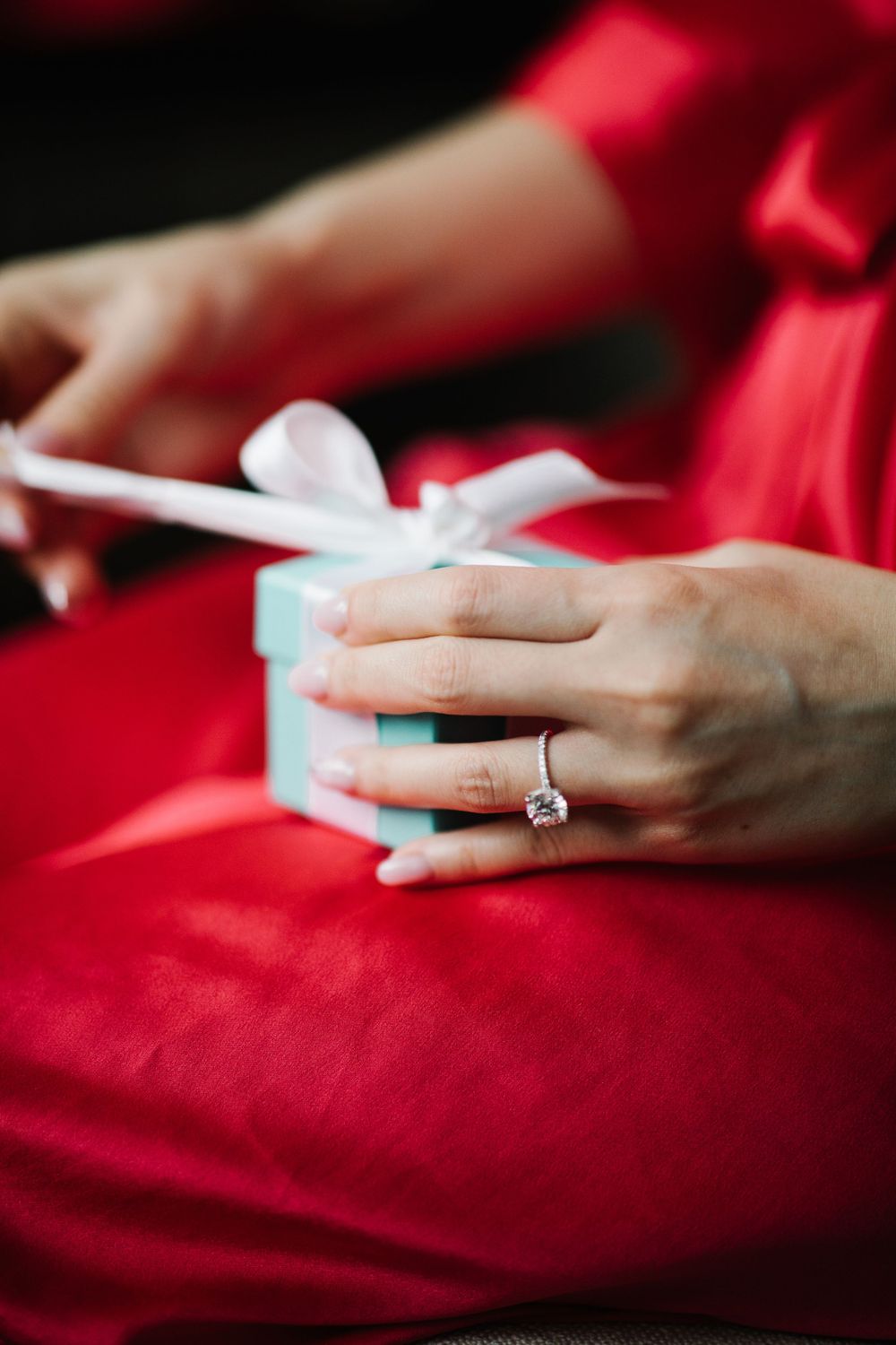 Hands holding small gift box with engagement ring against red fabric.