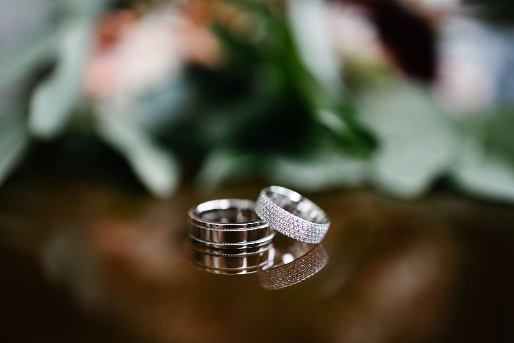 Two silver wedding rings with intricate details displayed on a dark surface with blurred floral background.
