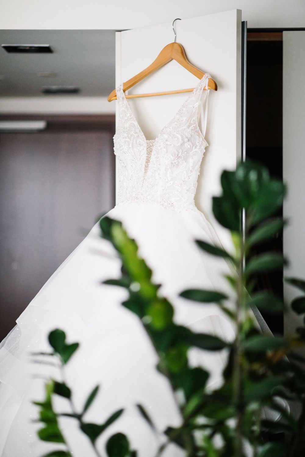 Elegant white wedding dress hanging on wooden hanger with green plants in foreground.