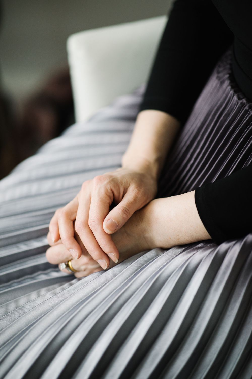 Hands with wedding ring resting on pleated grey fabric.