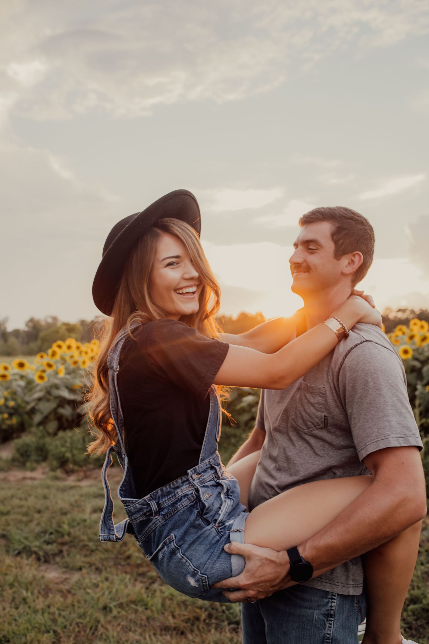 A romantic couple shares a playful moment in a sunflower field at sunset with golden light behind them.