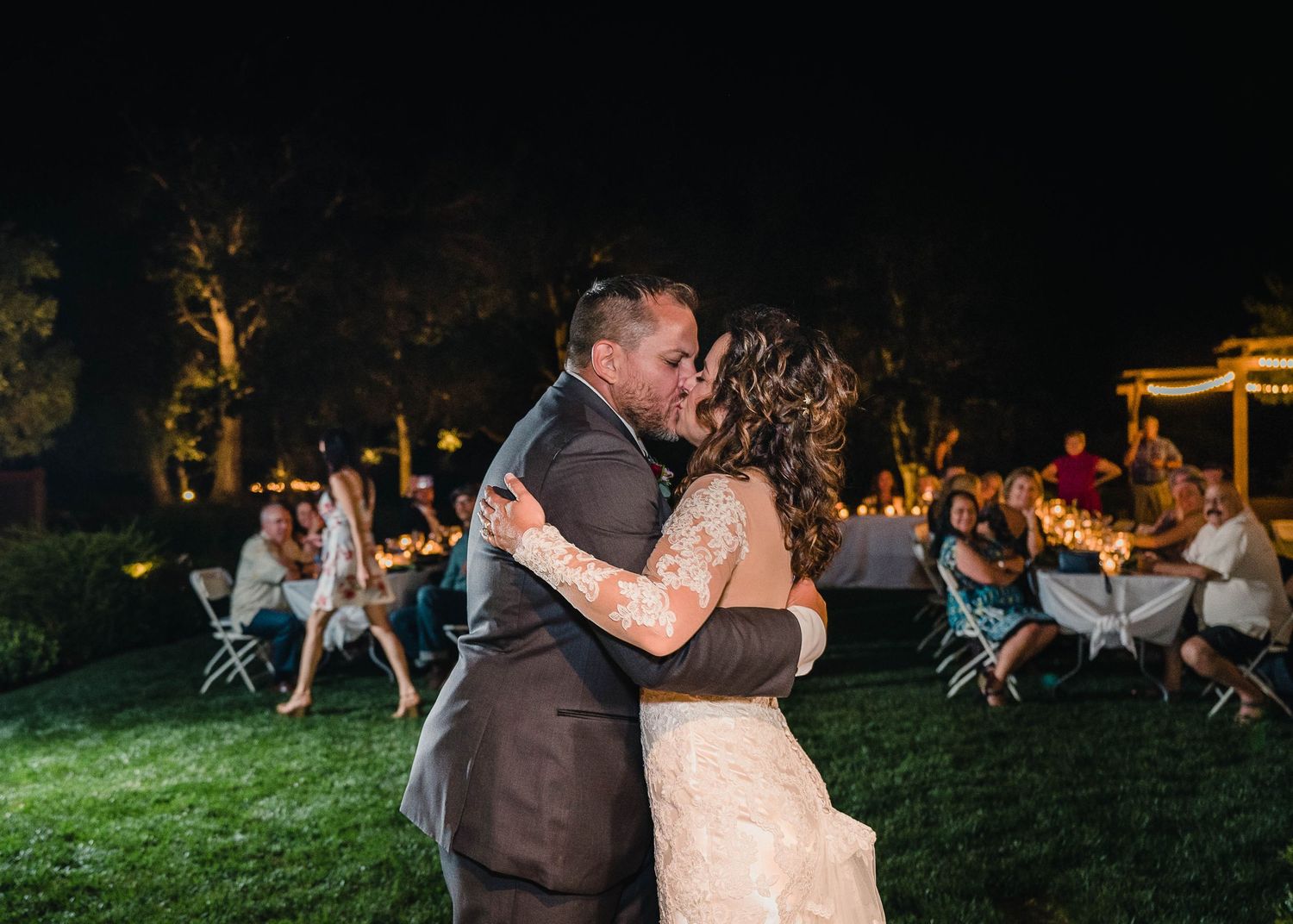 Couple embraces during outdoor evening wedding dance with guests seated at tables in background.