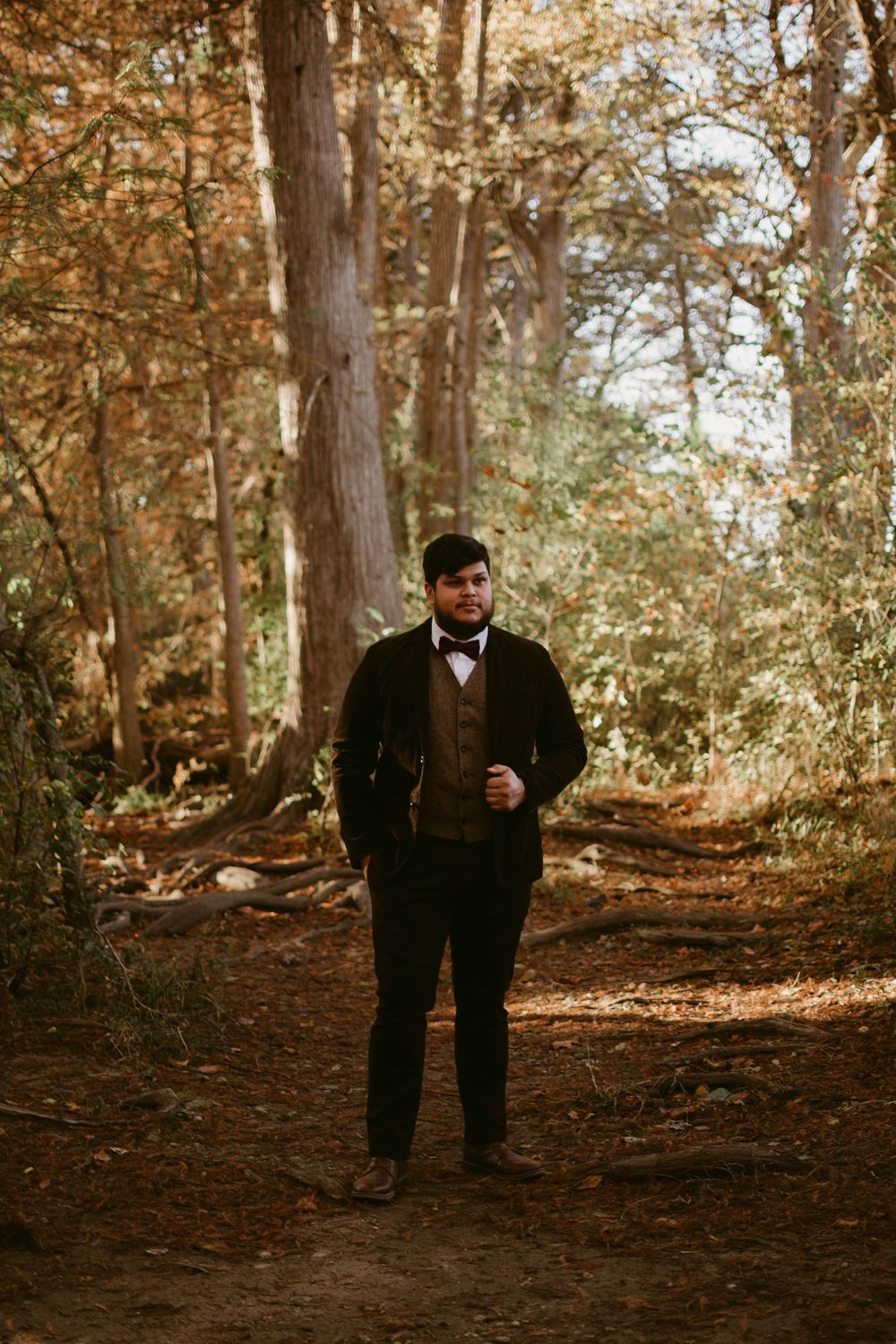 A full-length formal portrait of someone in a black suit standing on a forest path.