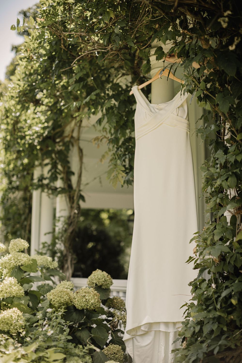 A white wedding dress hanging among green foliage and hydrangea bushes in natural lighting.