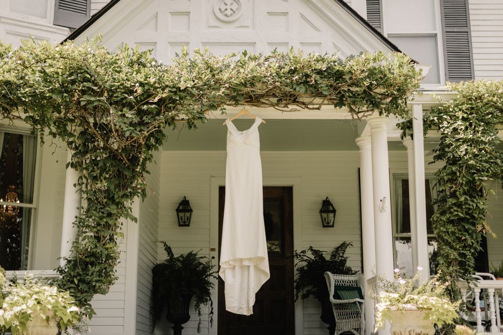 Elegant white wedding dress hanging on rustic porch with climbing vines and greenery.