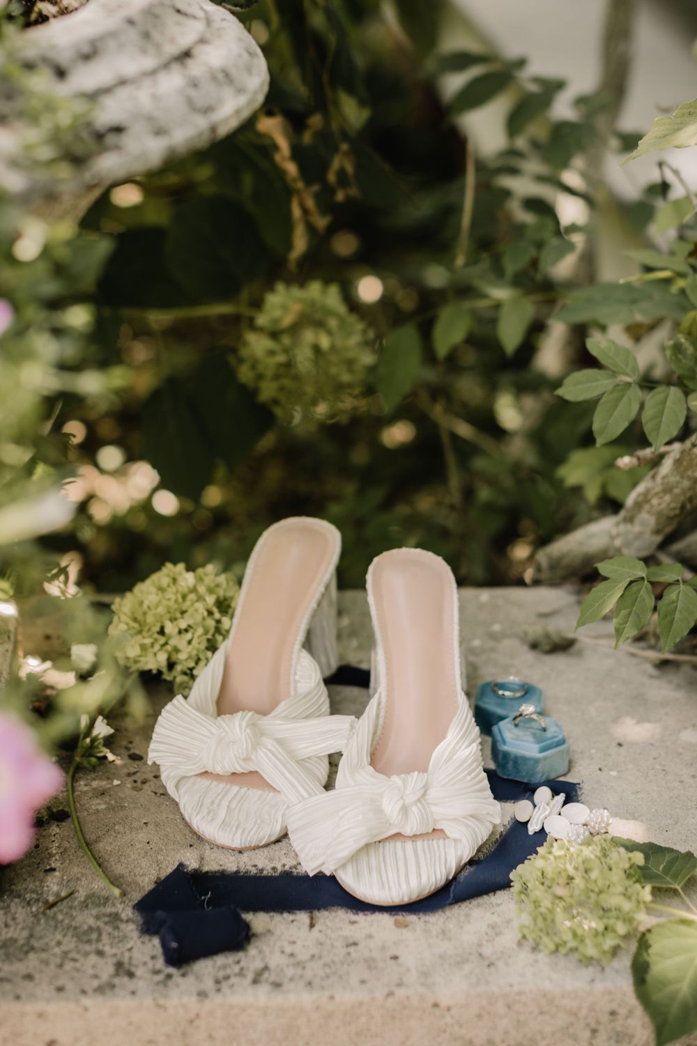 White bow-detailed bridal shoes displayed with blue accessories against a garden backdrop.