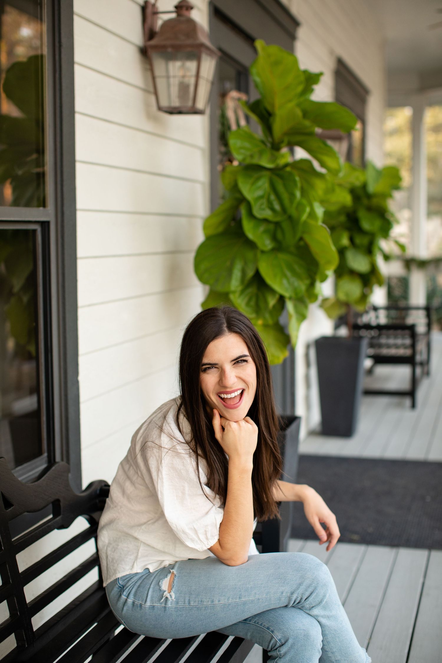 Woman in white shirt and jeans relaxes on porch bench near potted plants.