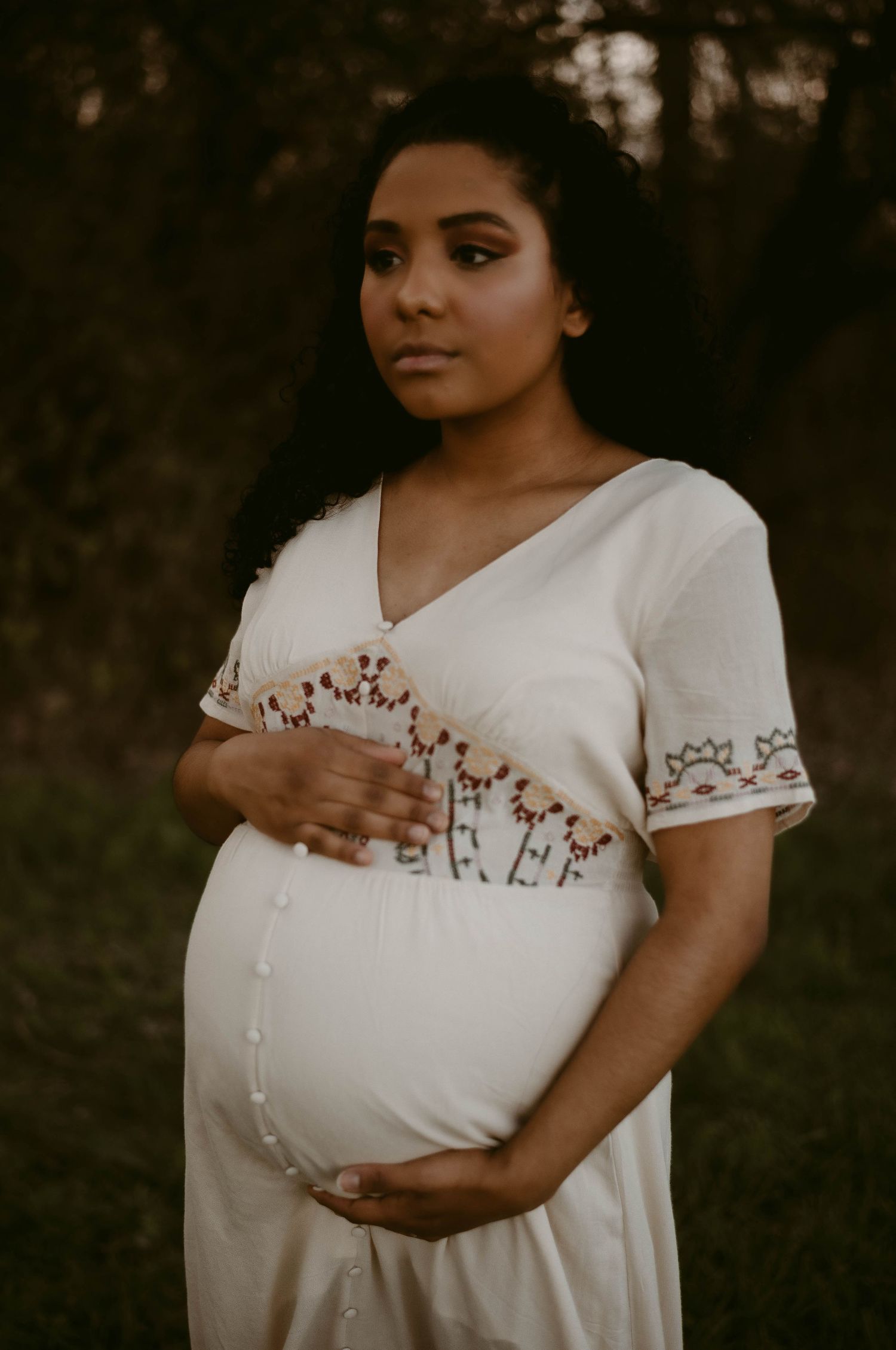 Maternity portrait in a white embroidered dress against a natural wooded backdrop.