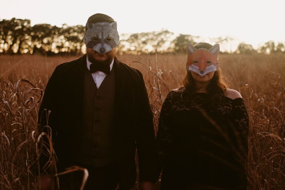 Two people wearing fox masks pose together in formal attire at sunset.