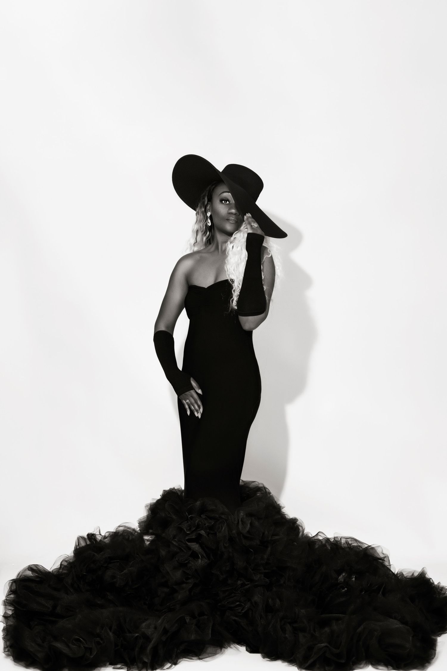 Glamorous black and white photo of a figure in a dramatic black gown and wide-brimmed hat.