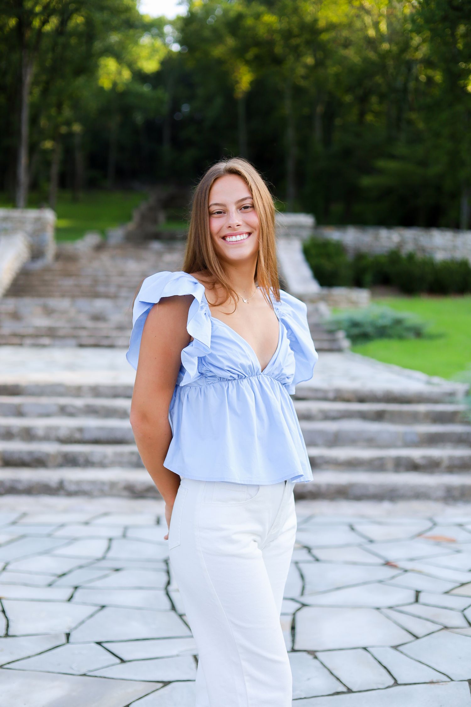A girl in a blue top and white jeans smiling in front of steps.