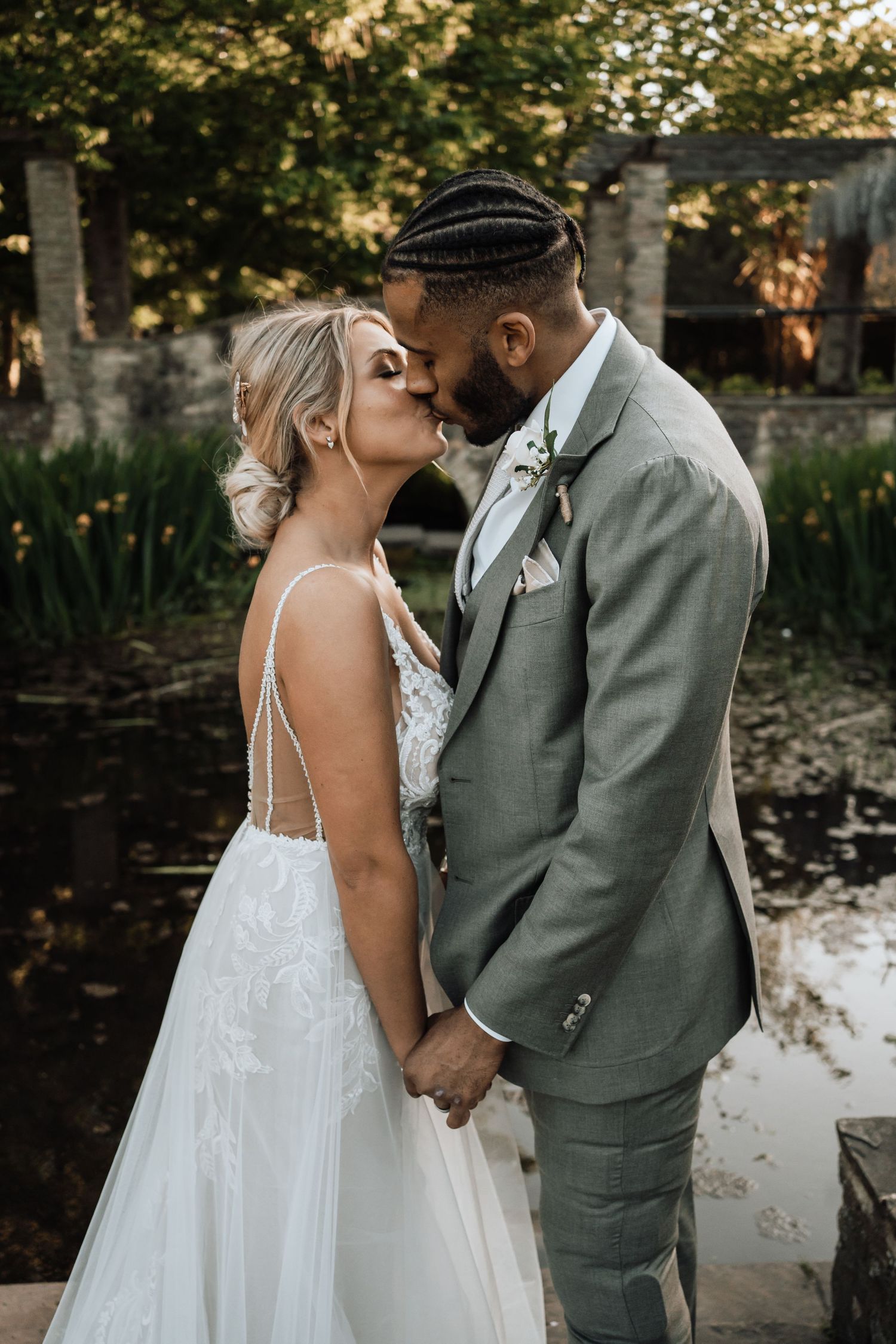 A bride and groom holding hands and kissing with greenery and a pond behind them.