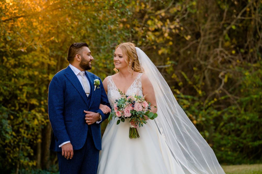 A bride and groom pose outdoors during autumn with fall foliage in the background.