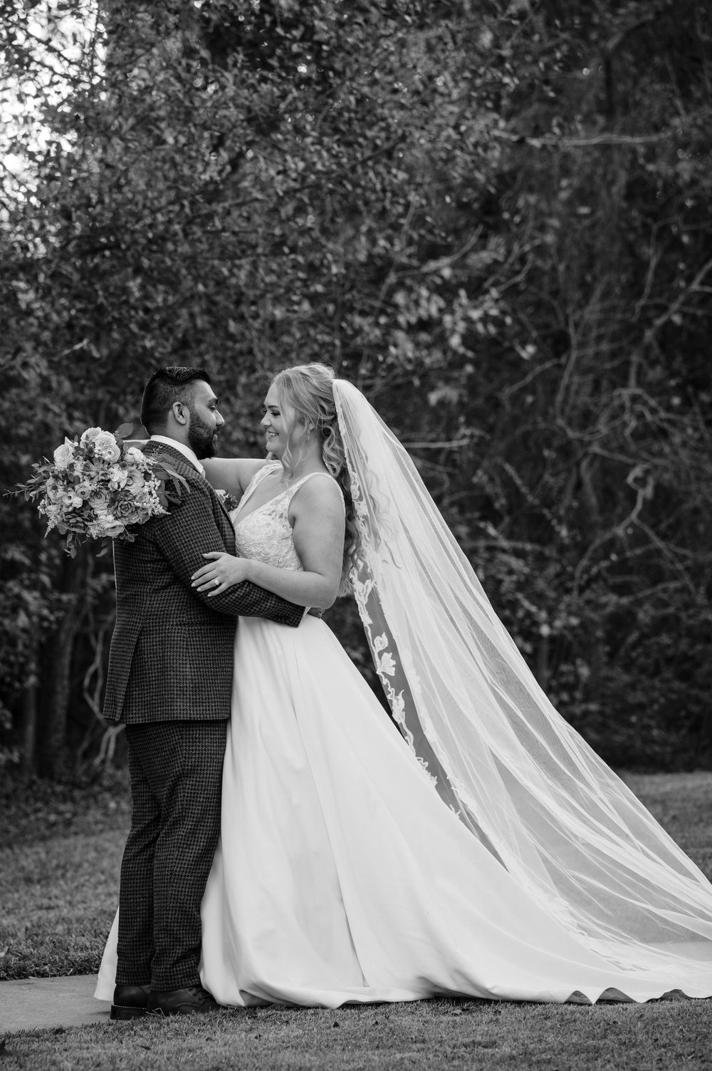 A dramatic black and white wedding portrait with a flowing veil in an outdoor setting.