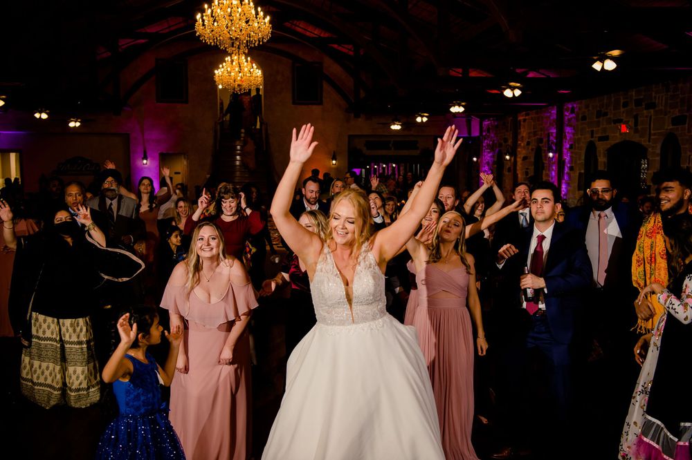 A bride celebrates on the dance floor surrounded by wedding guests in colorful attire.