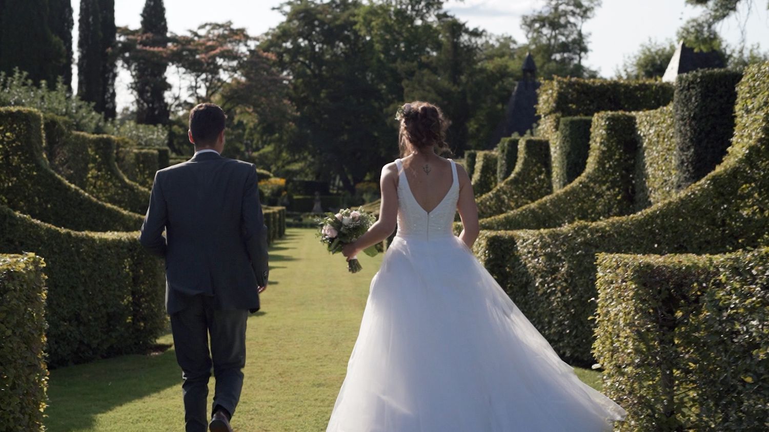 Wedding couple walks through a lush garden pathway lined with manicured hedges and trees.