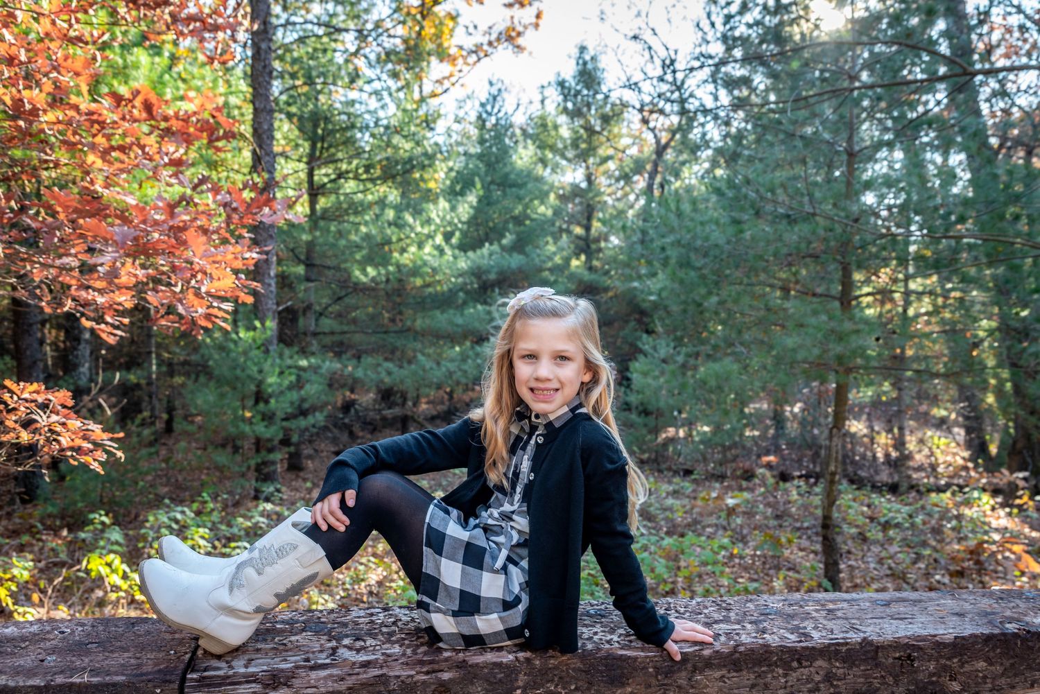 Child sitting on wooden log in autumn forest wearing plaid and black clothing.