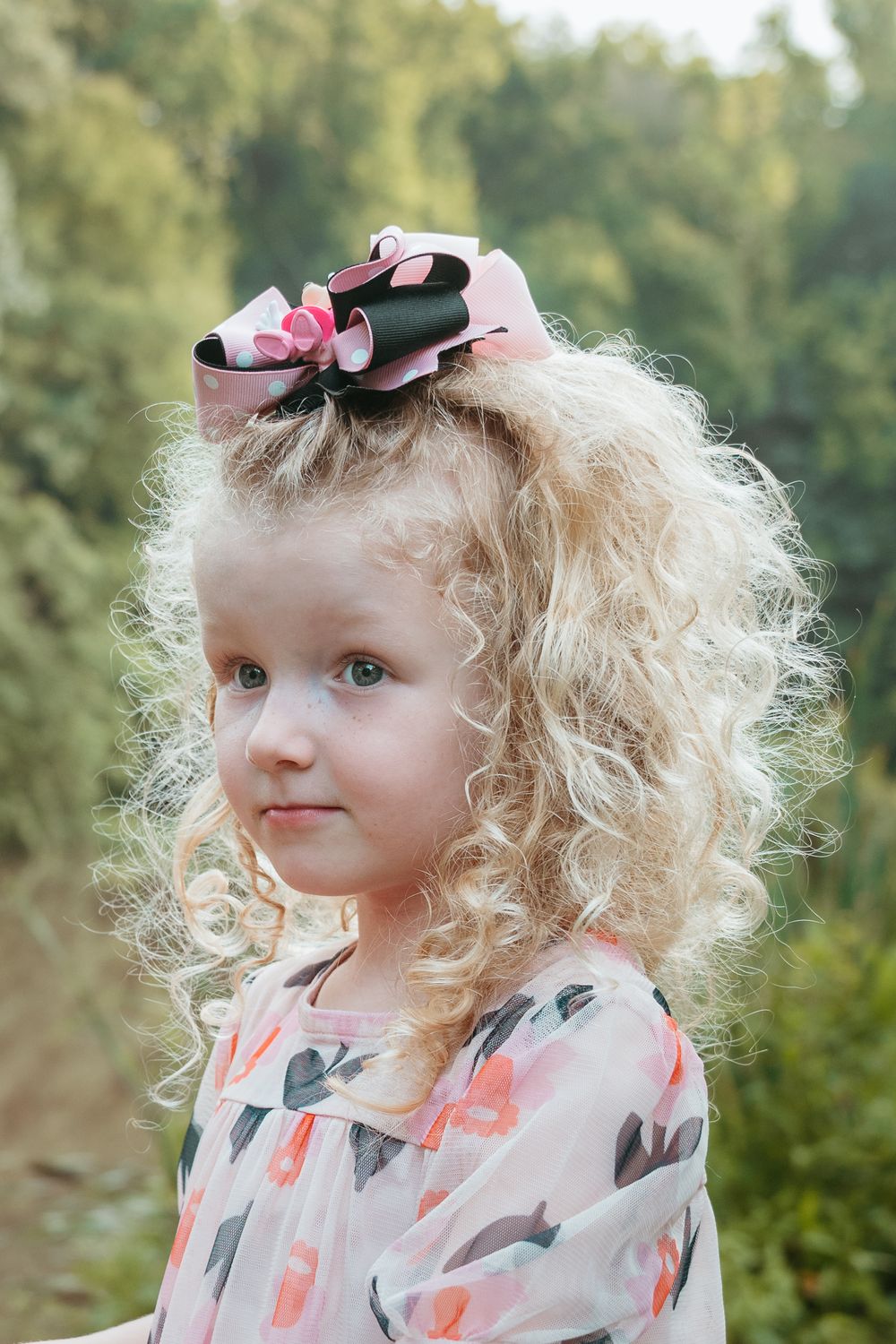 Young child with curly blonde hair wearing a floral dress and pink bow.