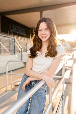 Casual dancer in white top and jeans leans on railing with warm sunset light creating a glowing backlit portrait.