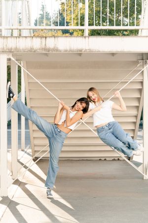 Two dancers wearing jeans pose playfully while holding white support beams in a bright outdoor corridor.