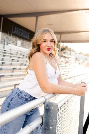 Casual dancer in white tank top and jeans leans against metal railing at outdoor stadium bleachers during sunset.