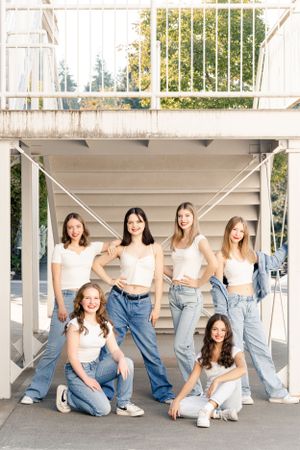 A group high school dancers in white tops and blue jeans pose together under a covered outdoor area in a coordinated casual style.