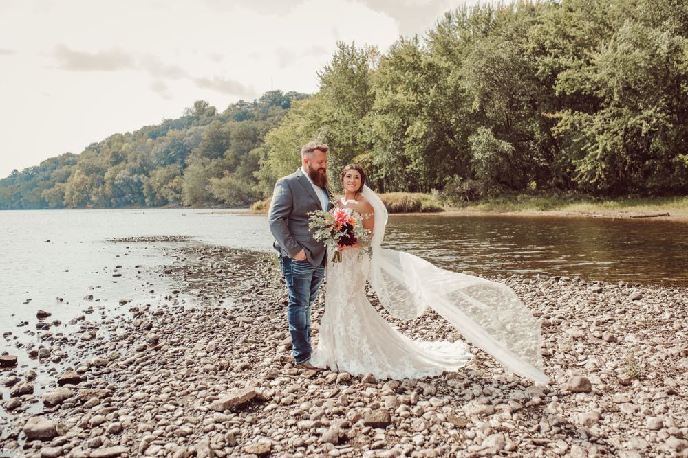Newlyweds pose on rocky shoreline with flowing wedding dress and lake backdrop.
