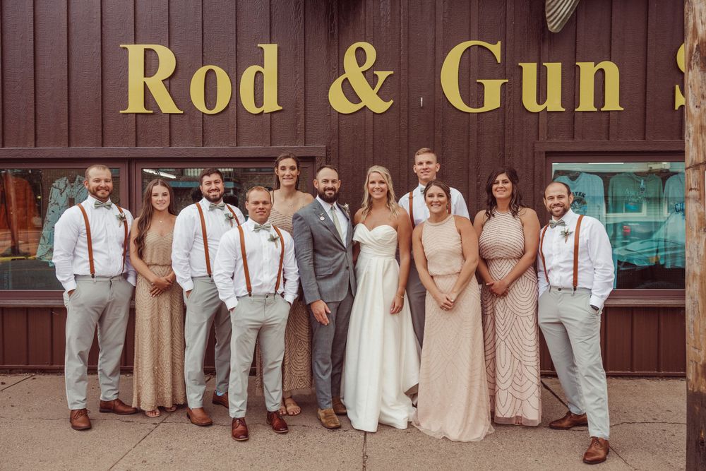 Wedding party poses in front of rustic Rod & Gun sign wearing coordinated beige and grey attire.