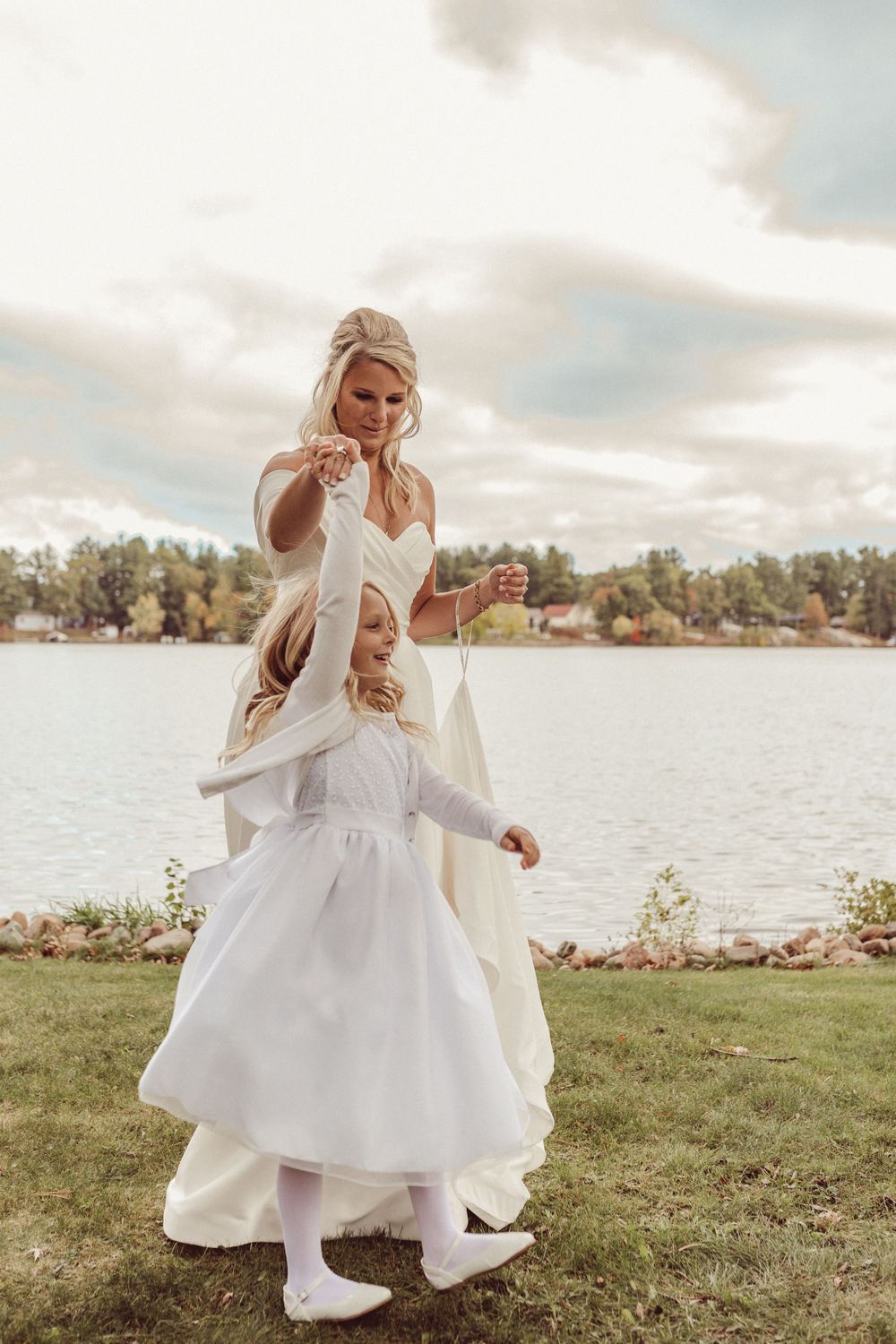 Person in white wedding dress dances playfully on grassy lakefront.