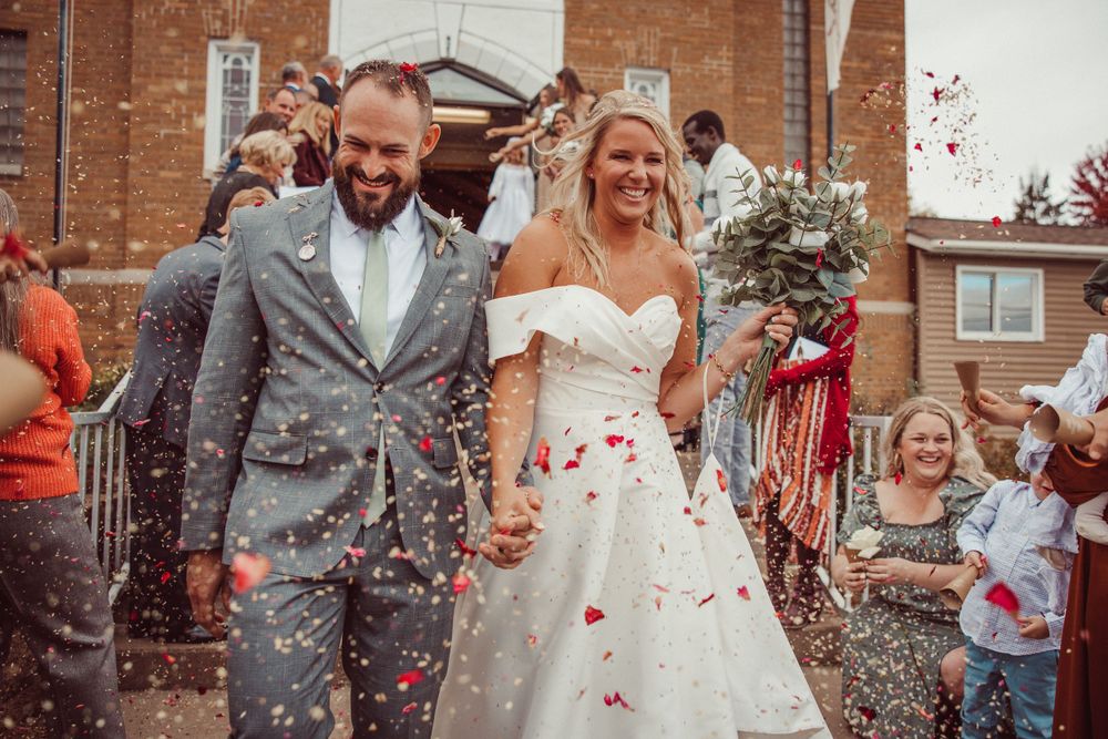 Newlyweds exit celebration amid shower of red confetti outside brick building.