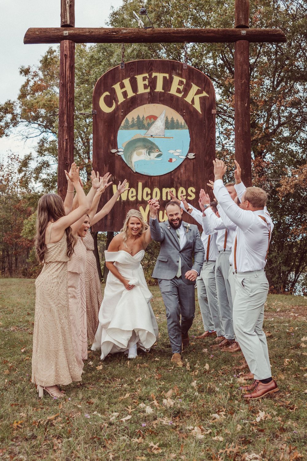 Wedding party celebrates under wooden Chetek welcome sign featuring fish and lake artwork.