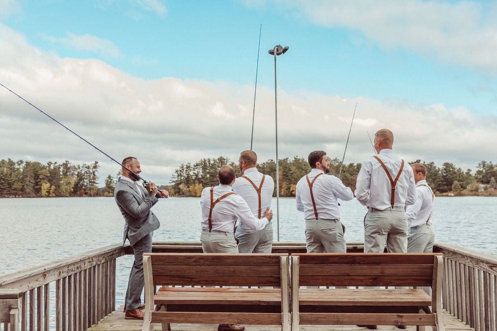 Groomsmen in suspenders fish from wooden dock overlooking peaceful lake.