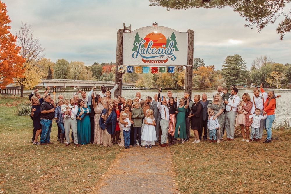 Large wedding party poses outdoors under vintage-style park entrance sign in autumn.