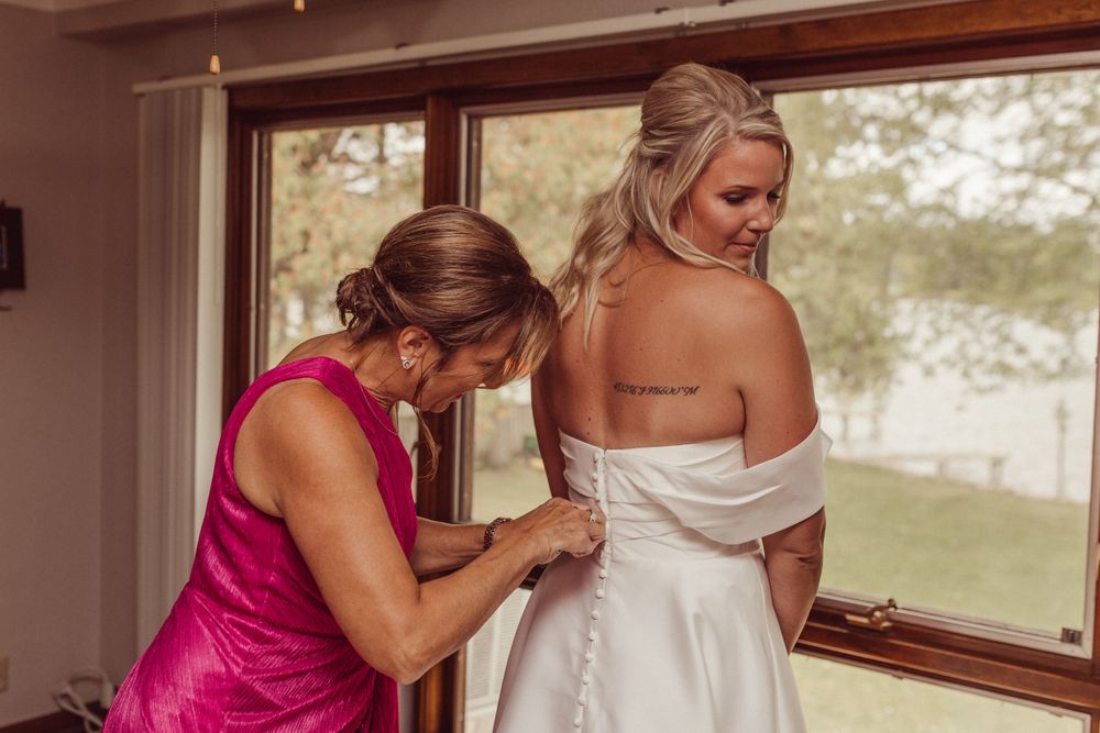 A woman in pink helps adjust the back of an elegant white wedding dress.