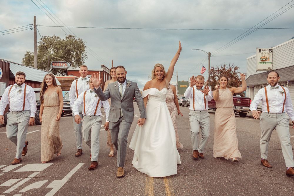 Wedding party walks down middle of small town street in coordinated neutral outfits.