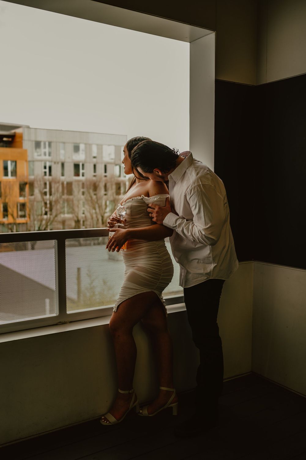 A couple sharing a tender moment by large windows overlooking city buildings.