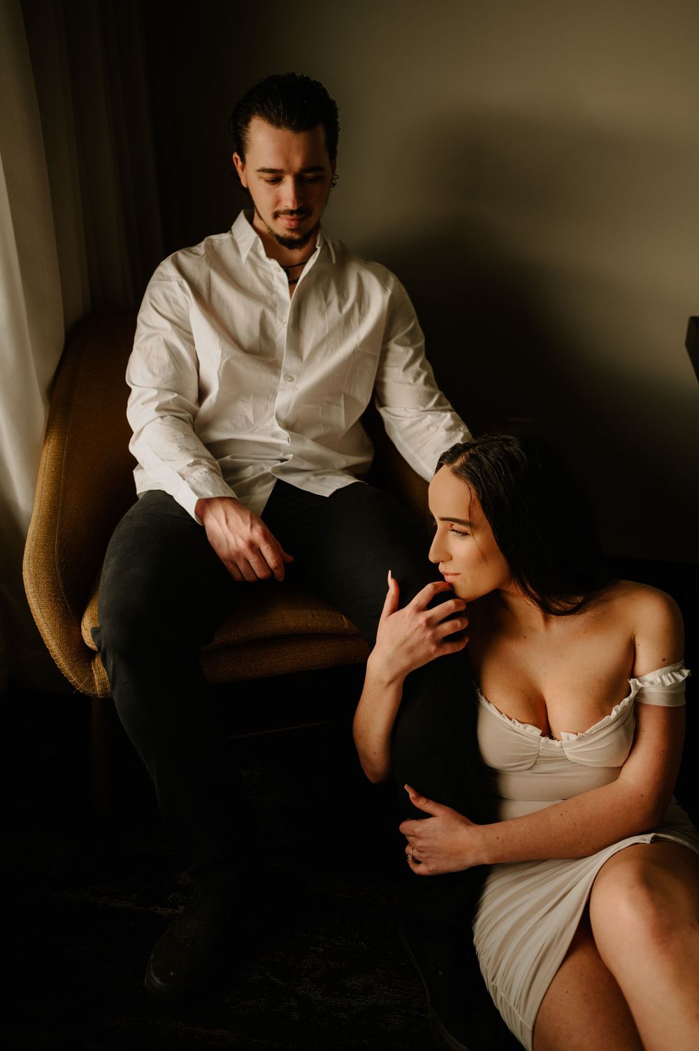 Intimate couple in moody lighting with white shirt and dress sitting on brown armchair.
