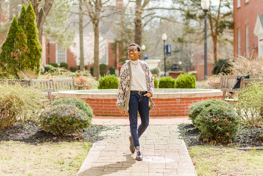 Walking portrait on campus pathway wearing cardigan and jeans among landscaped grounds.