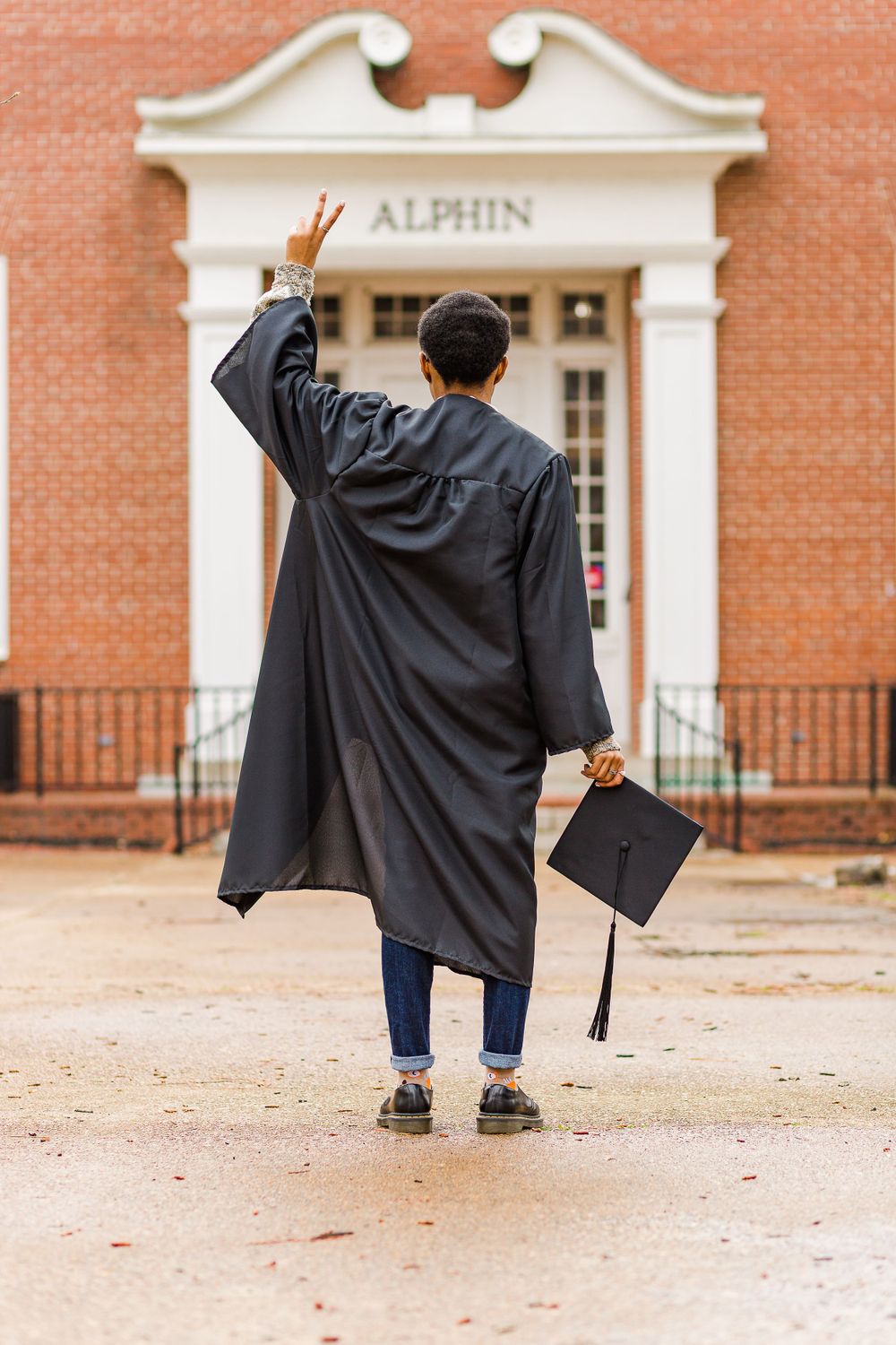 Graduate in black gown holds mortarboard while facing Alphin building entrance.