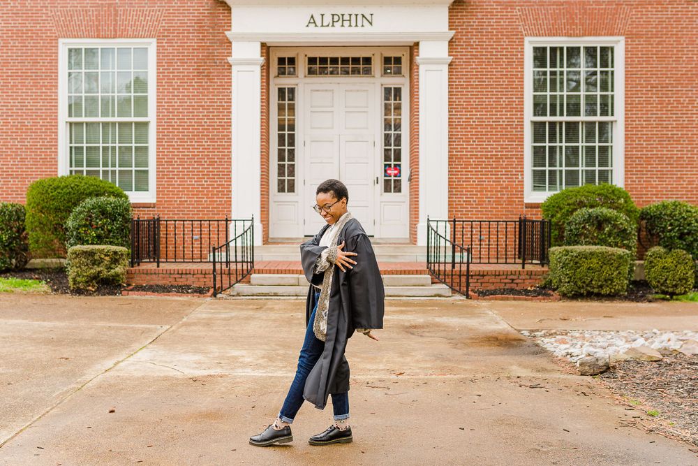 Graduate walks in front of red brick building with white columns and 'ALPHIN' signage.