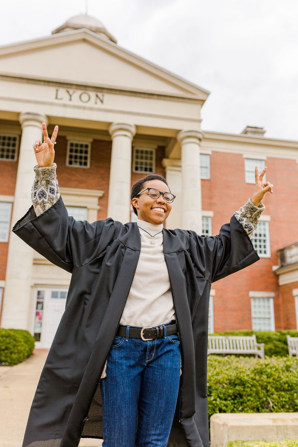 Graduate in academic regalia celebrates with victory signs in front of Lyon building.