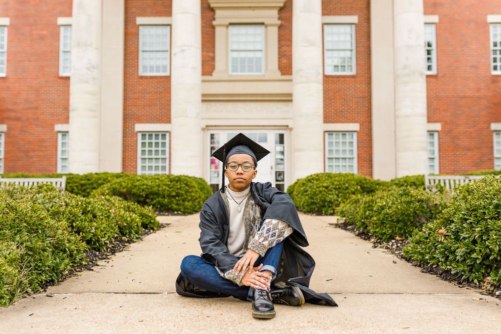 Graduate in cap and gown sits on pathway leading to red brick academic building.