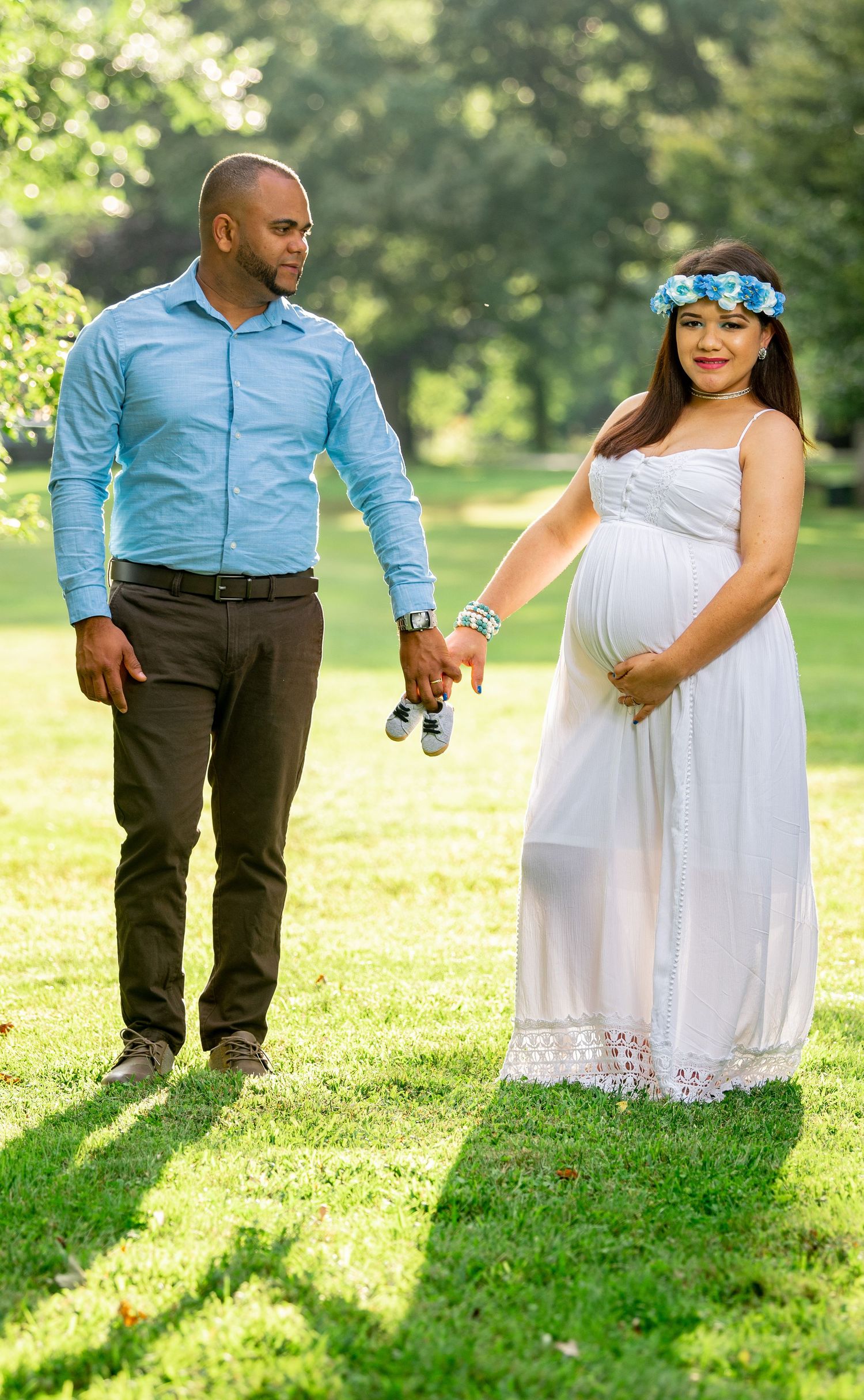 A couple holds hands in a sunny garden setting during their maternity photoshoot.