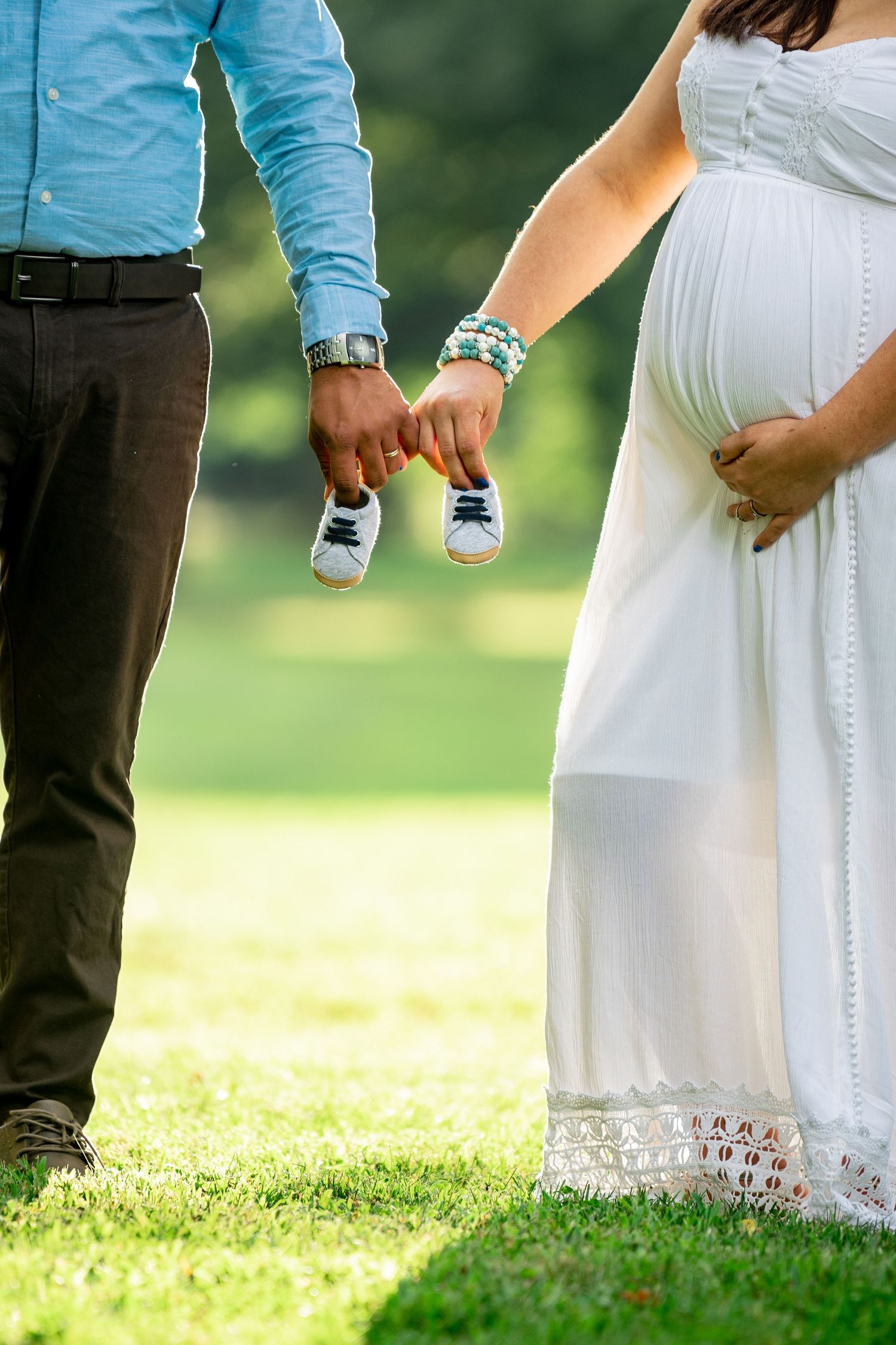 Couple holds tiny baby shoes while showcasing pregnancy in white dress outdoors.