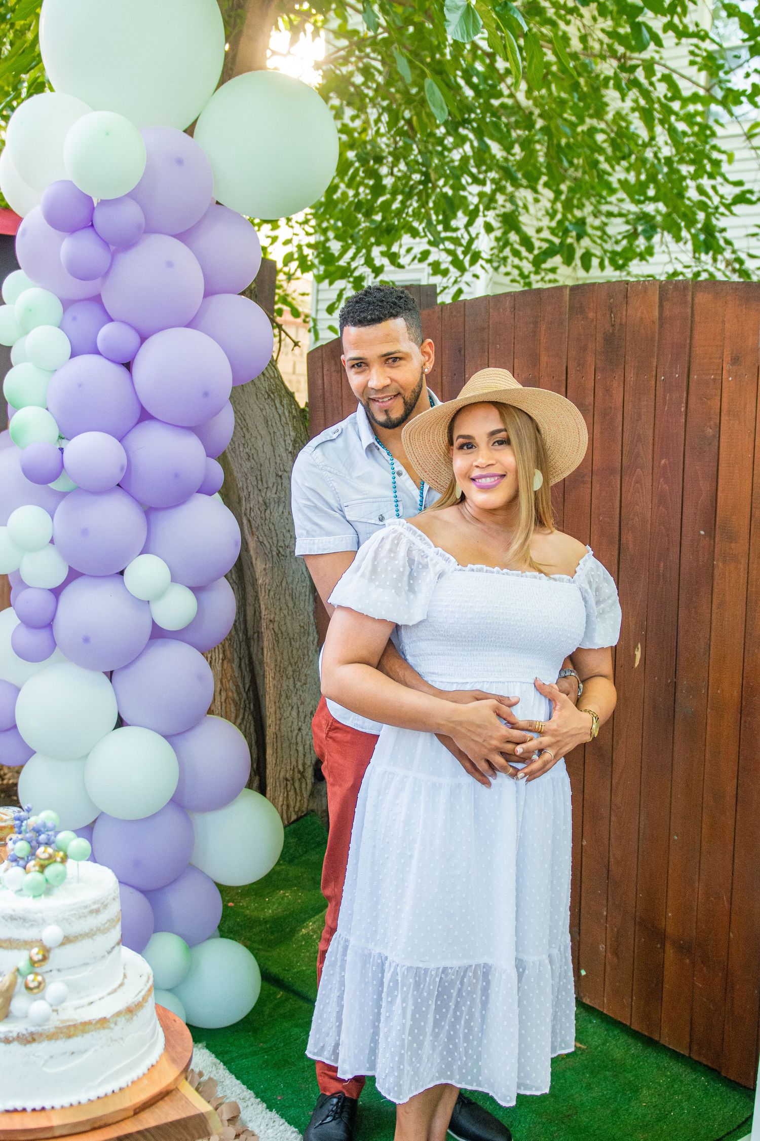 Backyard celebration with purple and white balloon arch, tiered cake display on artificial grass.