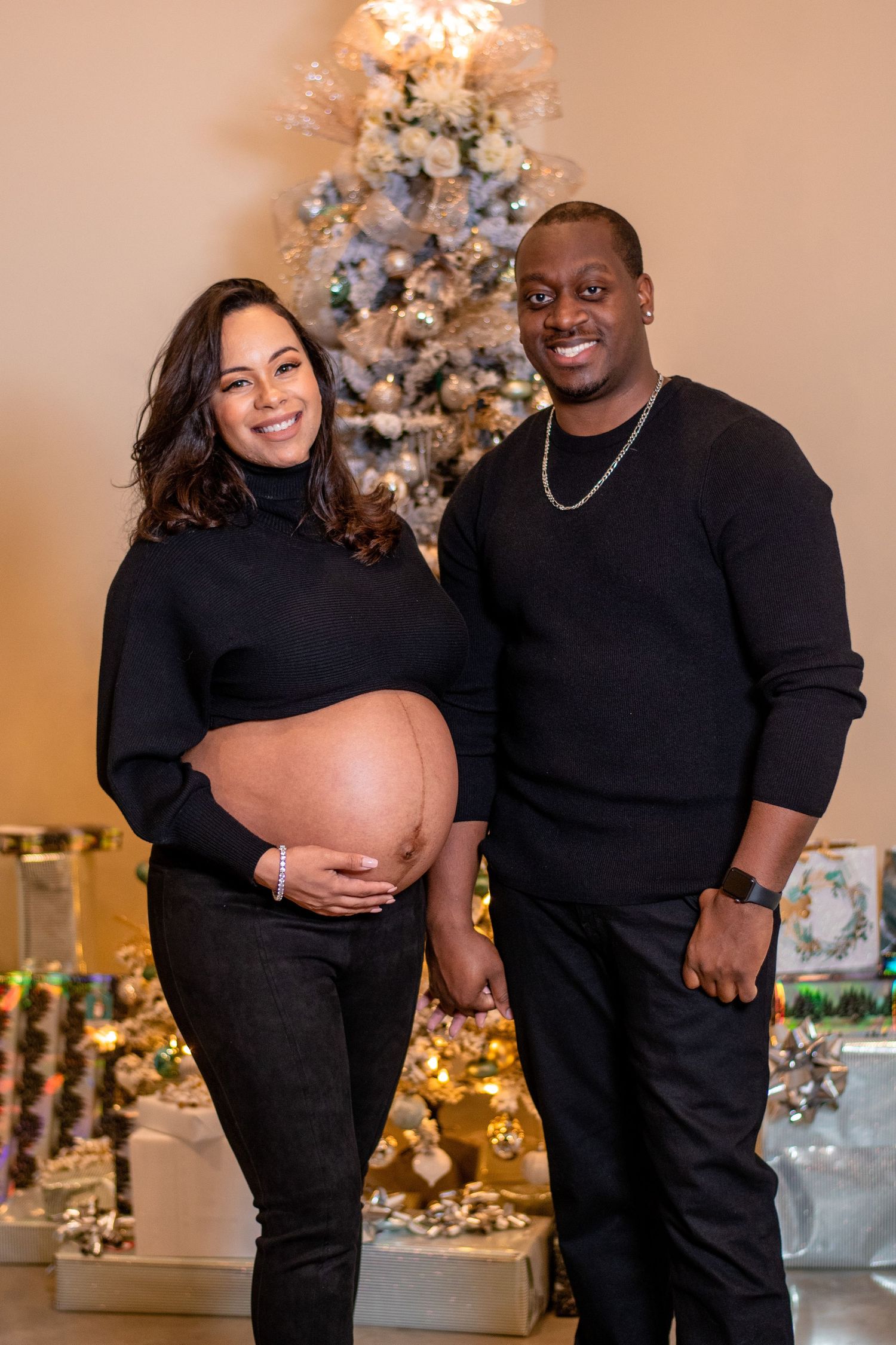 A couple poses in matching black outfits in front of a decorated Christmas tree.