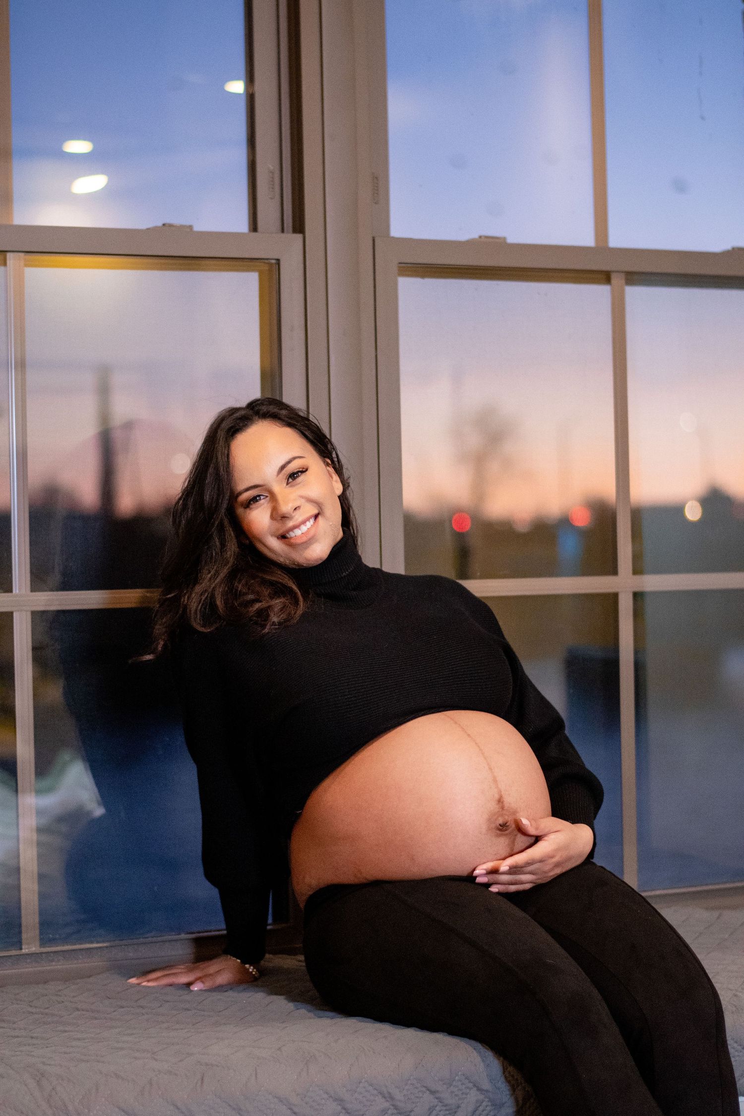 A pregnant woman in black lounges by a window during sunset for a maternity photo.