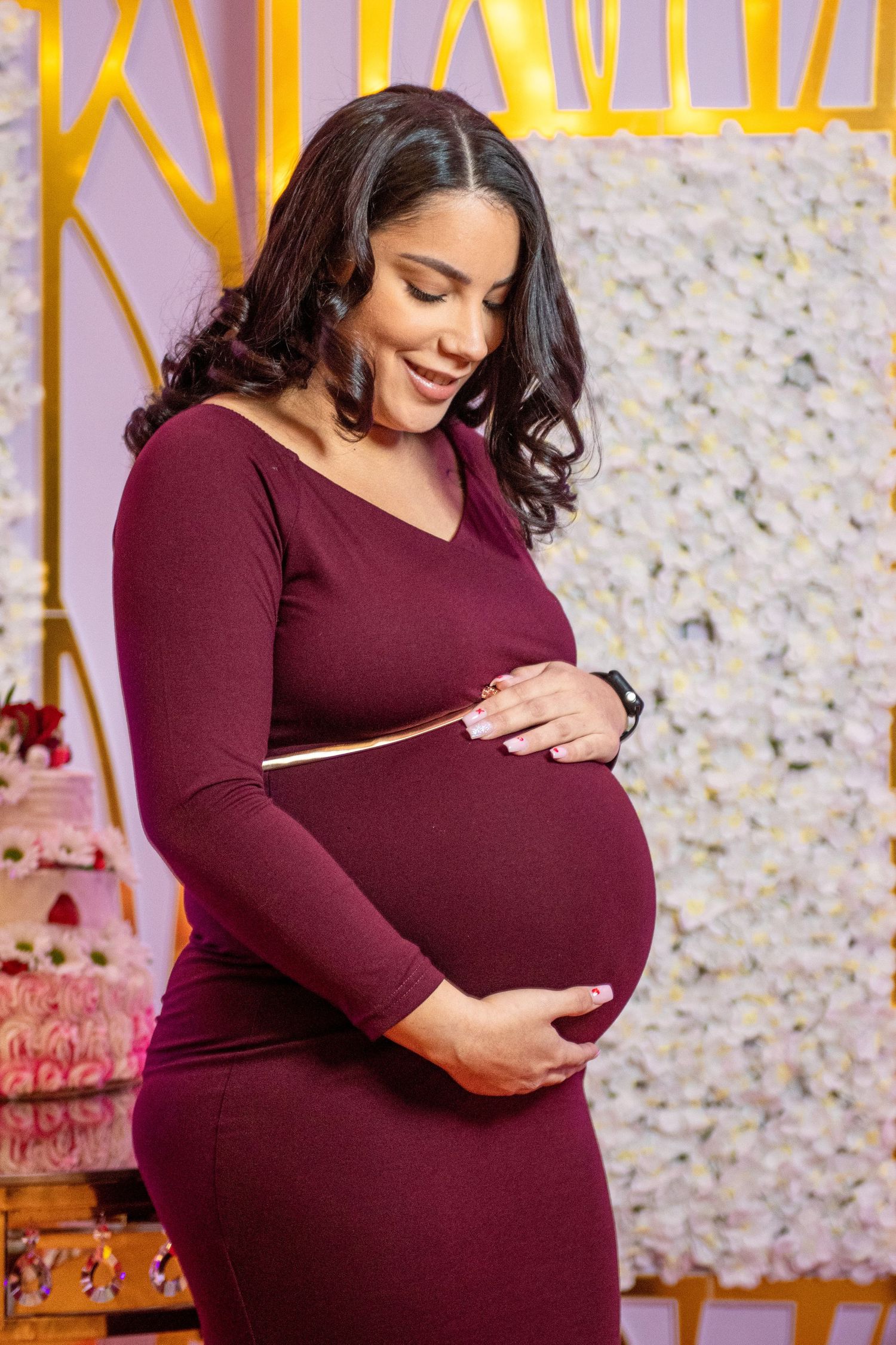 A pregnant woman in burgundy dress touches her belly against a decorative gold and white backdrop.