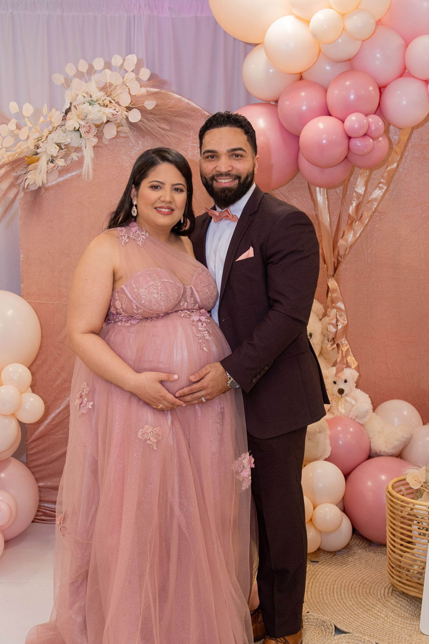Maternity photo featuring a couple in formal attire against a pink balloon backdrop with floral decor.