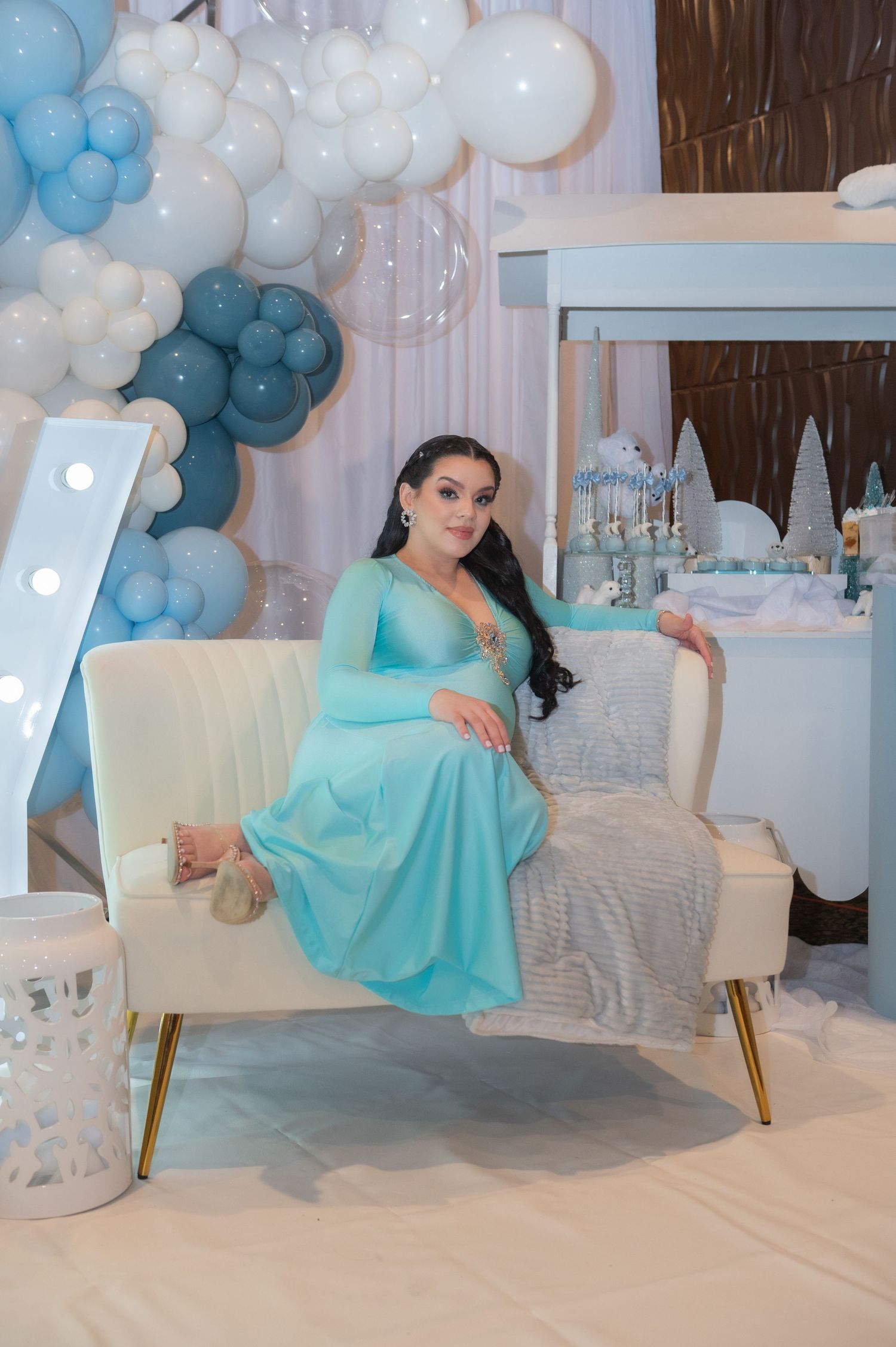 Woman in turquoise gown poses on white chair with blue and white balloon arch decor.